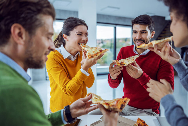 Pizzazeit. Eine aufgeregte multikulturelle Gruppe von Geschäftsleuten steht im Büro und isst mittags Pizza. Teambildung in der Büropause. Die lächelnde Frau hatte gerade einen Bissen von der Pizza genommen - Foto, Bild