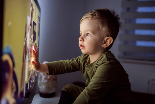 Children's addiction to television and cartoons. A boy touches the TV screen with one hand. Close up of a kid sitting right in front of the TV in his pajamas and staring at a cartoon. Modern parenting - Photo, Image
