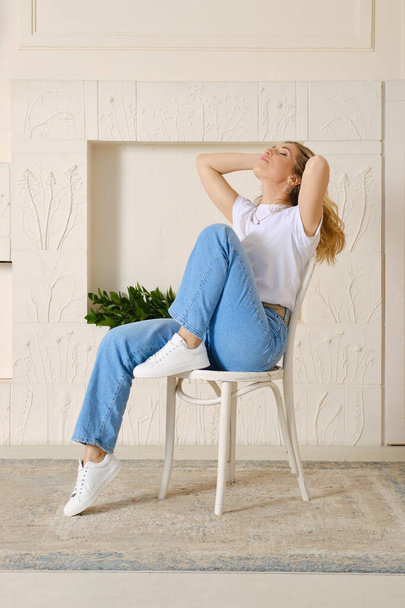 Relaxed woman in wide jeans and t-shirt sits on chair and throws her head back - 写真・画像