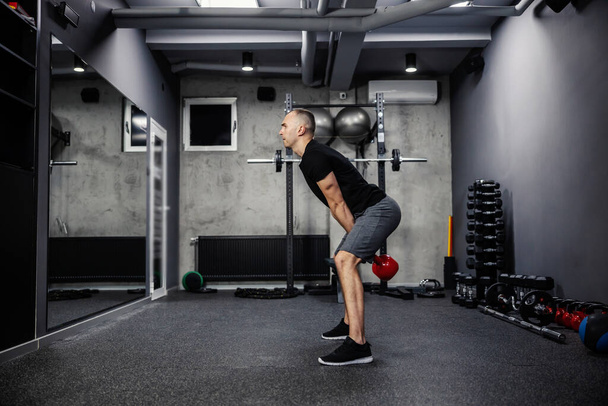 Dead lifting workout with a kettle bell. Man in sportswear with a black T-shirt stands with his legs spread and lifts a kettle bell with both arms in gym. Strength and physical endurance - Fotoğraf, Görsel