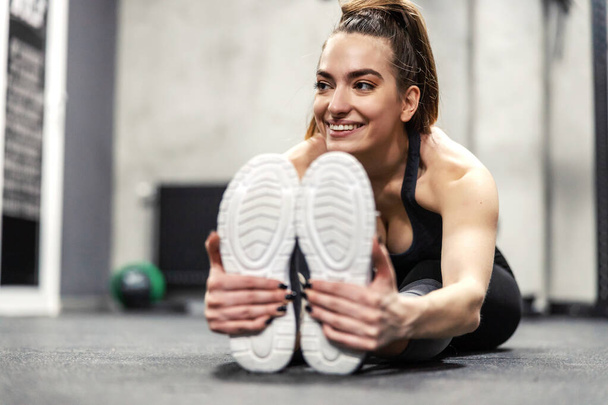 Muscle stretching in spot training. A female person sits on the gym floor and stretches her leg and foot muscles. Women's hands hold the sole of the sneaker and stretch the leg muscles, worming up - Photo, Image