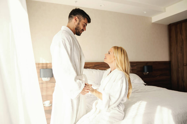 Love attraction and respect. Waking up in a warm hotel with natural light. A man stands above the bed and gently holds the girl's hands. She sits on the bed, they both have white bathrobes on. Loving - Photo, Image