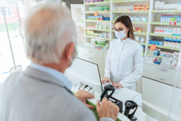 Buying and selling prescription drugs and pharmacist advice. An adult female pharmacist standing behind the counter and selling drugs to a mature man. She is wearing a protective mask - Photo, Image