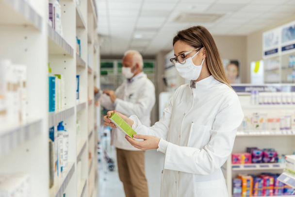 Egy női gyógyszerész áll egy gyógyszeres polc mellett a koronavírus idején. Arcmaszkot és fehér egyenruhát visel. Egy csomag gyógyszert tart a kezében, és felolvassa a nyilatkozatot. - Fotó, kép