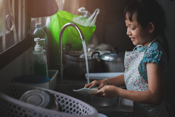 Ασιατικό κοριτσάκι πλένει πιάτα στην κουζίνα με χαμόγελο και χαρούμενη - Φωτογραφία, εικόνα