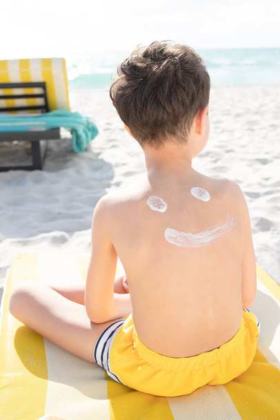Op het strand wordt zonnebrandcrème aangebracht op de rug van het kind, grappige smiley gezicht is geschilderd met zonnebrandcrème. Concept van bescherming tegen de zon op het strand, lotion beroertes, preventie van zonnebrand. Tan. Zomer. - Foto, afbeelding