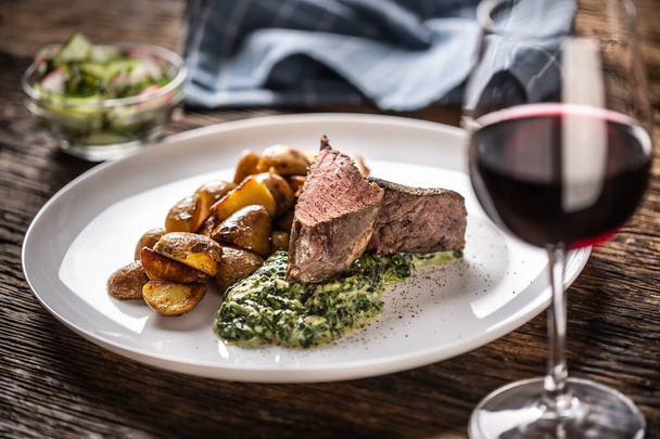 Red wine as a typical baverage to a red meat sous vide steak with roasted potatoes, spinach dip and fresh salad served on a white porcelain plate on the wooden desk. - Фото, зображення
