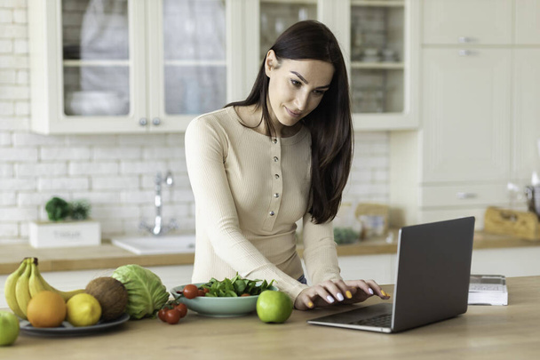 Szczęśliwy kaukaska kobieta stoją w kuchni domowej z laptopem i zestaw zdrowej żywności, wygląda w Internecie do przepisów kulinarnych do gotowania żywności dietetycznej - Zdjęcie, obraz