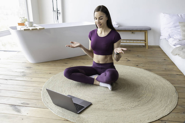 Young woman fitness trainer in sportswear is sitting on the floor with laptop at home. Online sports concept - Foto, afbeelding