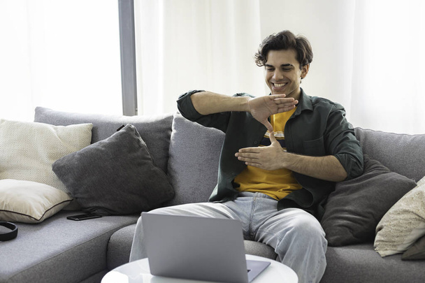 Deaf or hard hearing happy smiling young caucasian man uses sign language while video call using laptop while sitting on the couch at home - 写真・画像