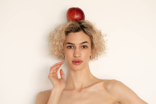 portrait of a young beautiful woman with an apple on her head, stands near a white wall - Foto, Imagem