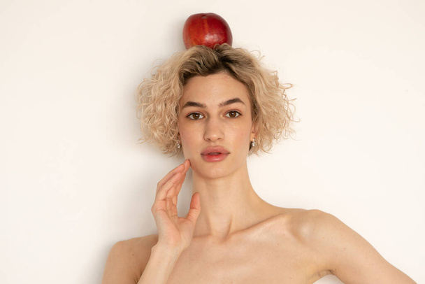portrait of a young beautiful woman with an apple on her head, stands near a white wall - Photo, Image