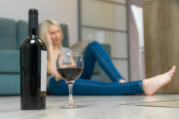 Une jeune femme blonde est assise sur le sol près du canapé et tenant sa tête, au premier plan se trouve une bouteille de vin à moitié vide et un verre de vin. - Photo, image