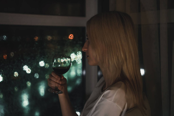 Nuoren vaalean naisen muotokuva valkoisessa paidassa punaviinilasillisen kanssa katselemassa yökaupunkia ikkunan läpi. Valikoiva fokus - Valokuva, kuva