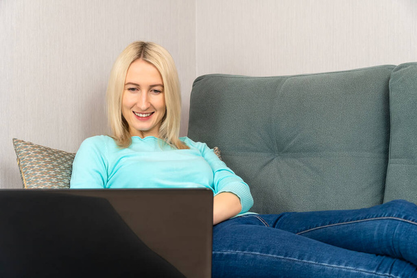 Giovane bella donna bionda con un sorriso utilizza un computer portatile mentre si trova sul divano a casa - Foto, immagini