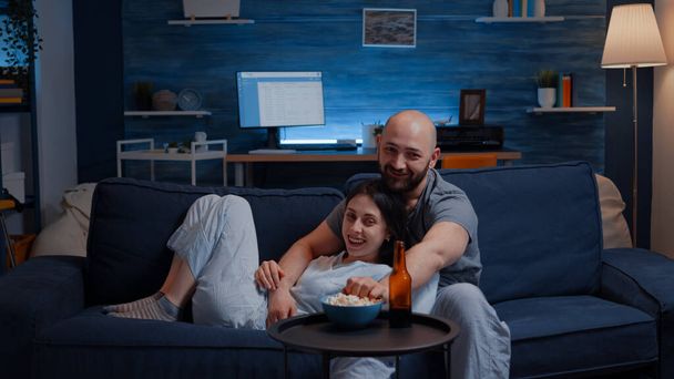 Όμορφο ευτυχισμένο ζευγάρι βλέποντας τηλεόραση στον καναπέ χαλαρώνοντας το βράδυ γελώντας - Φωτογραφία, εικόνα