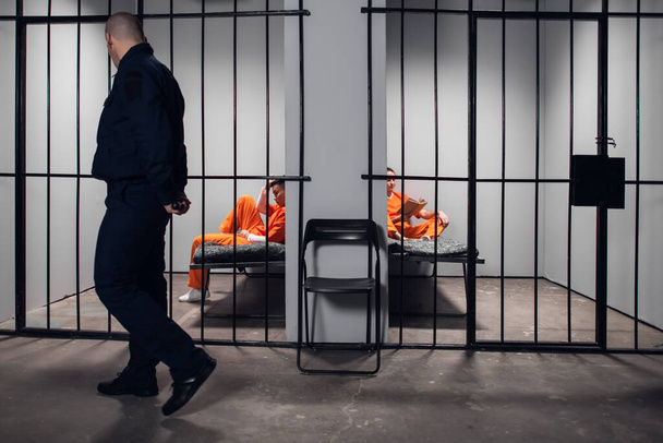 Een gevangenisbewaker met een wapenstok maakt een rondleiding door de gevangeniscellen voordat het licht uitgaat. Twee gevangenen in rode gewaden zitten achter de tralies. - Foto, afbeelding