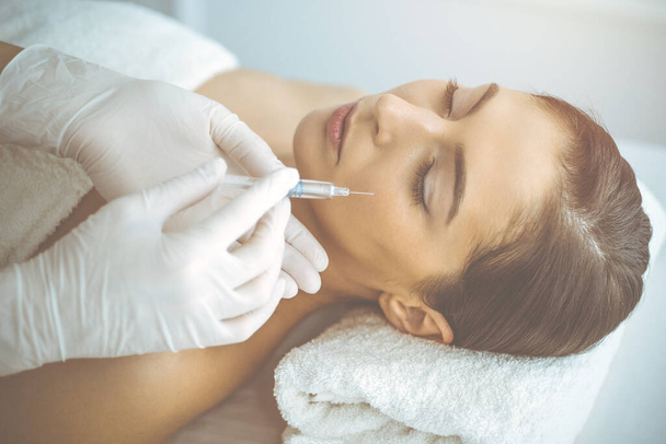 Beautician doing beauty procedure with syringe to face of young brunette woman in sunny clinic. Cosmetic medicine and surgery, beauty injections - Foto, Imagem