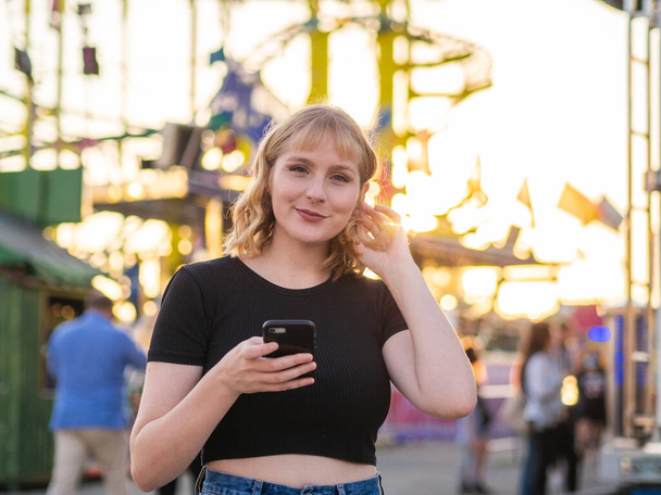 A Spanish blonde woman holding a phone near a rollercoaster in the park during the sunset - Photo, Image