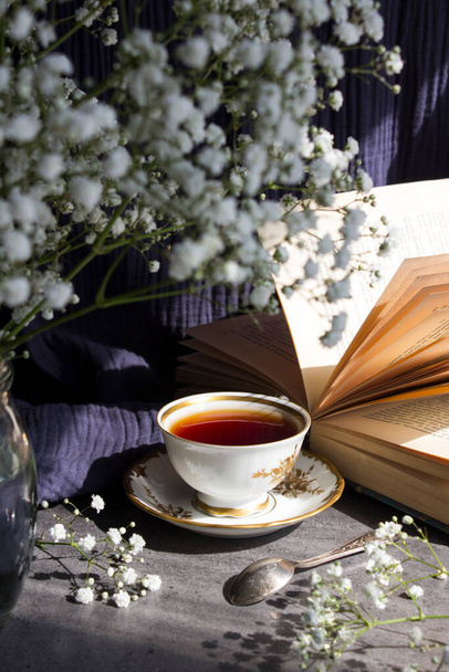 Натюрморт з чашкою чаю. Проста композиція фото з маленькими білими квітами і чашкою чорного чаю на столі. Прекрасне ранкове зображення
.  - Фото, зображення