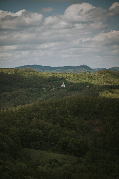 A vertical shot of thick trees covering hills beneath a cloudy sky with a small church standing out - Foto, imagen