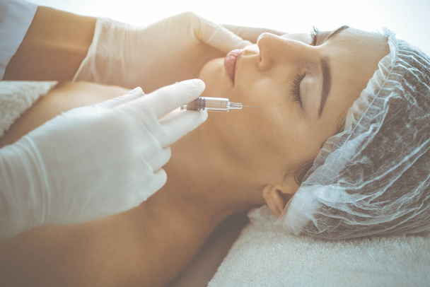 Beautician doing beauty procedure with syringe to face of young brunette woman in sunny clinic. Cosmetic medicine and surgery, beauty injections - Foto, immagini