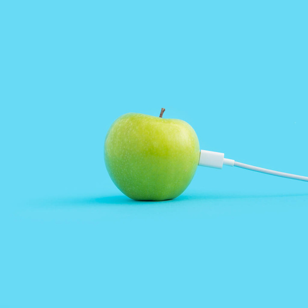 A white phone charger attached to a green apple isolated on a blue background - Valokuva, kuva