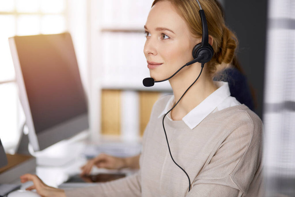 Businesswoman talking by headset while sitting in sunny office. Call center and diverse people group in business - Fotoğraf, Görsel