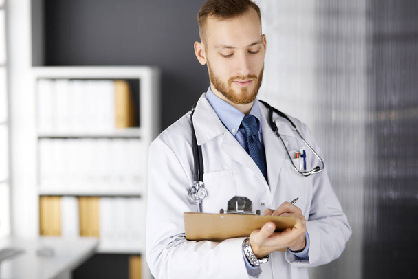 Дружній червонобородатий лікар стоїть і пише з буфера в клініці на своєму робочому місці. Ідеальна медична служба в лікарні. Медицина, охорона здоров'я
 - Фото, зображення