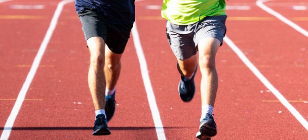 Close up das pernas de dois meninos do ensino médio correndo lado a lado em pistas em uma faixa vermelha em luz do sol brilhante. - Foto, Imagem