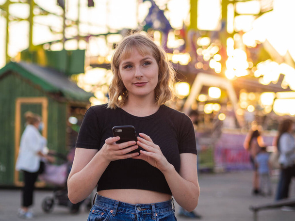 A Spanish blonde woman holding a phone near a rollercoaster in the park during the sunset - Foto, afbeelding