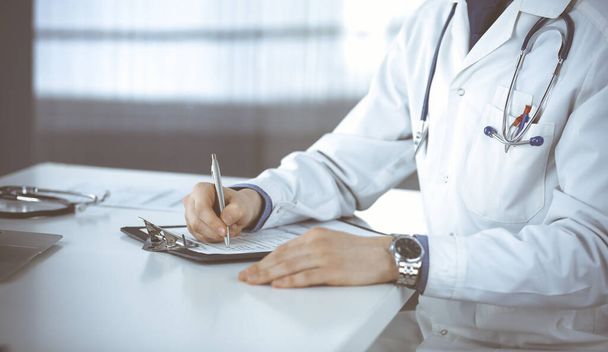 Unknown male doctor sitting and working with clipboard of medication history record in clinic at his working place, close-up. Young physician at work. Perfect medical service, medicine concept - Foto, Imagem