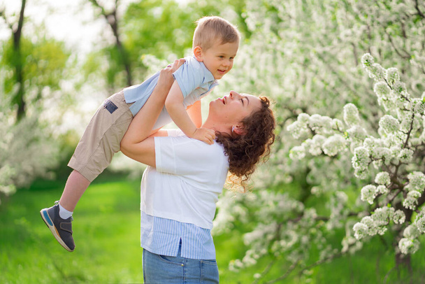 Mama bawi się w wiosennym ogrodzie z młodym synem, rzuca dziecko w górę.  - Zdjęcie, obraz