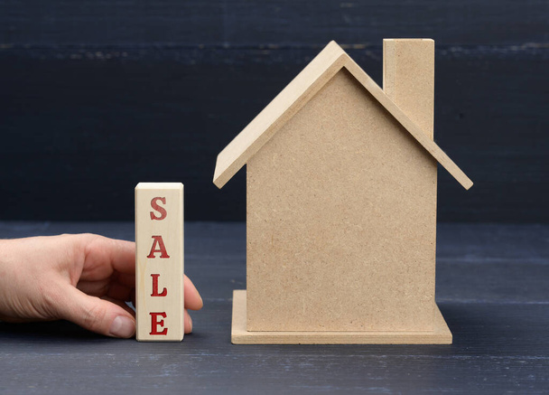 houten model van een huis en een hand houdt een blok met een inscriptie verkoop op een blauwe achtergrond. Huisverkoopconcept, vastgoedinvestering, makelaarsdiensten - Foto, afbeelding