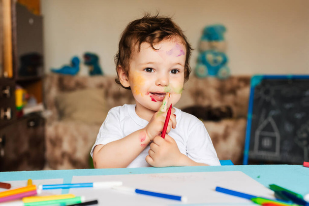 Ένα χαρούμενο χαρούμενο παιδί ζωγραφίζει με ένα στυλό άκρη τσόχας σε ένα άλμπουμ χρησιμοποιώντας μια ποικιλία εργαλείων σχεδίασης. - Φωτογραφία, εικόνα