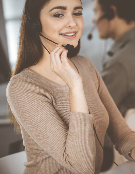 Young friendly girl in headsets is talking to a firms client, while sitting at the desk in a modern office together with her colleague. Call center operators at work - Φωτογραφία, εικόνα