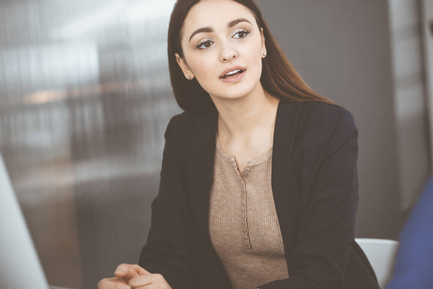 Young beautiful business woman is talking to her colleague, while sitting at the desk in a modern office. Concept of business success - 写真・画像