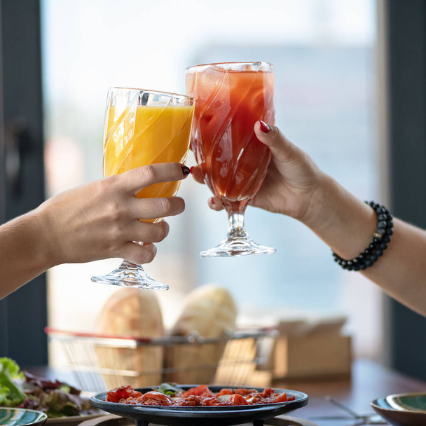 Festive toast. Female friends clink glasses with fresh juice or soft drink against the window on a blurred background. Close up shot. Bright colors of nature - Fotoğraf, Görsel