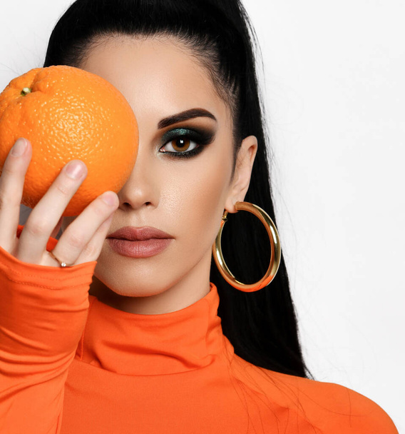 Young brunette woman in modern orange turtleneck sweater, shirt and earrings holds an orange in front of her face - Foto, imagen