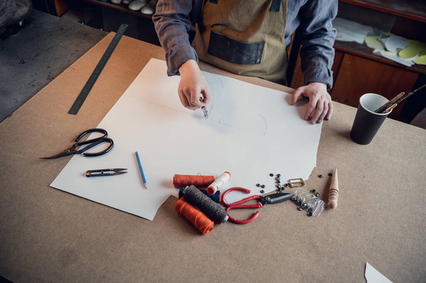 Un zapatero joven hace un dibujo para un patrón de zapatos de cuero en una mesa en su taller - Foto, imagen