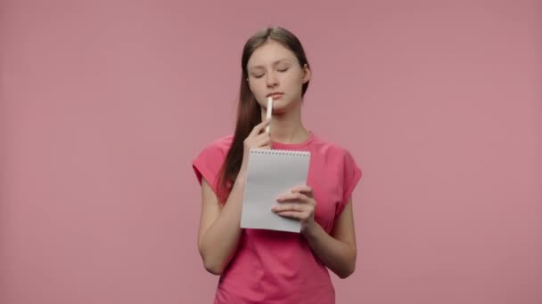 Portrait of fashion model is thinking and writing down her ideas in notebook. Young brown haired woman in pink t-shirt poses on pink studio background. Close up. Slow motion ready 59.94fps - Πλάνα, βίντεο