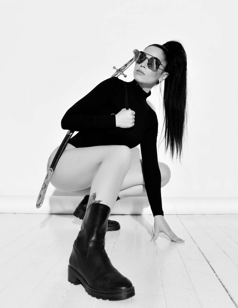 Black and white of young woman in bodysuit, brutal shoes and sunglasses sitting squatted, holding with sabre behind back - Foto, Imagen
