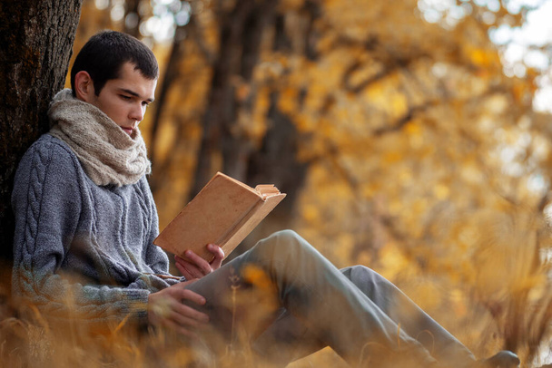 Egy fiatal férfi meleg kötött pulóverben és sálban Snood egy régi könyvet olvas az őszi parkban. Életmód - Fotó, kép
