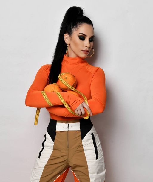 Young sporty slim woman in bright orange sportswear and turtleneck stands holding fresh oranges and measuring tape - Zdjęcie, obraz