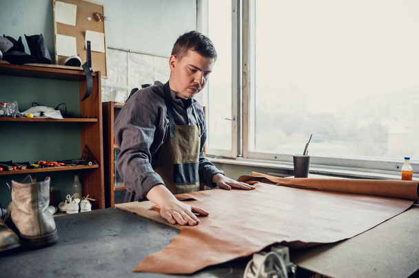 Um jovem aprendiz em uma oficina de inicialização prepara couro para uso posterior em uma mesa grande - Foto, Imagem