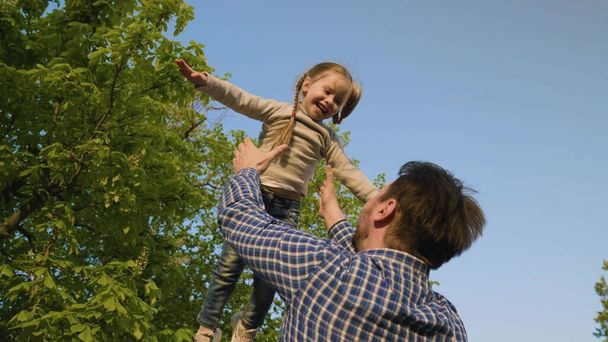 Baba mutlu kızını parktaki mavi gökyüzüne fırlatıyor, çocuk gülüyor ve seviniyor. Baba ve küçük çocuk birlikte oynuyor, gülüyor ve sarılıyorlar. Bebek ebeveynin kollarında. Babamla hafta sonu. Mutlu bir aile. - Fotoğraf, Görsel