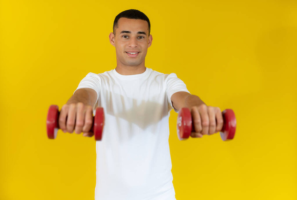 smiling handsome sportsman in white t-shirt holding dumbbell isolated over yellow background. - Photo, Image