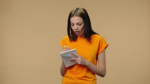 Portrait of fashion model thinking with concentration and writing thoughts and ideas in notebook. Young girl in yellow t-shirt posing on brown studio background. Close up. Slow motion ready 59.94fps. - Πλάνα, βίντεο