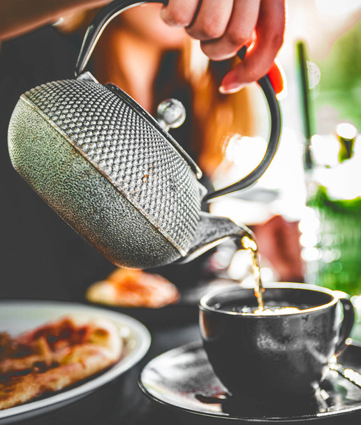 vrouw hand met metalen theepot giet zwarte thee in zwart glas kopje in cafe - Foto, afbeelding