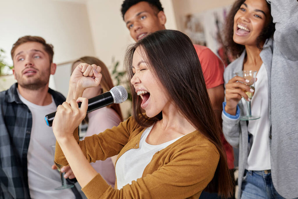 Leistung wie ein Superstar. Hübsches junges asiatisches Mädchen hält Mikrofon in der Hand und singt, während sie mit besten Freunden zu Hause Karaoke spielt, eine Gruppe von Leuten trinkt und zusammen Spaß hat. Selektiver Fokus - Foto, Bild